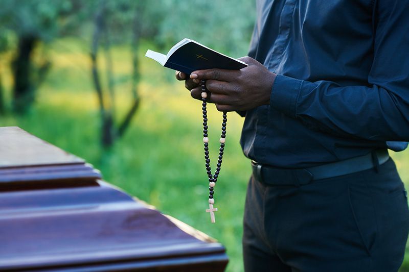 A man in a suit is holding a white rose in front of a coffin.