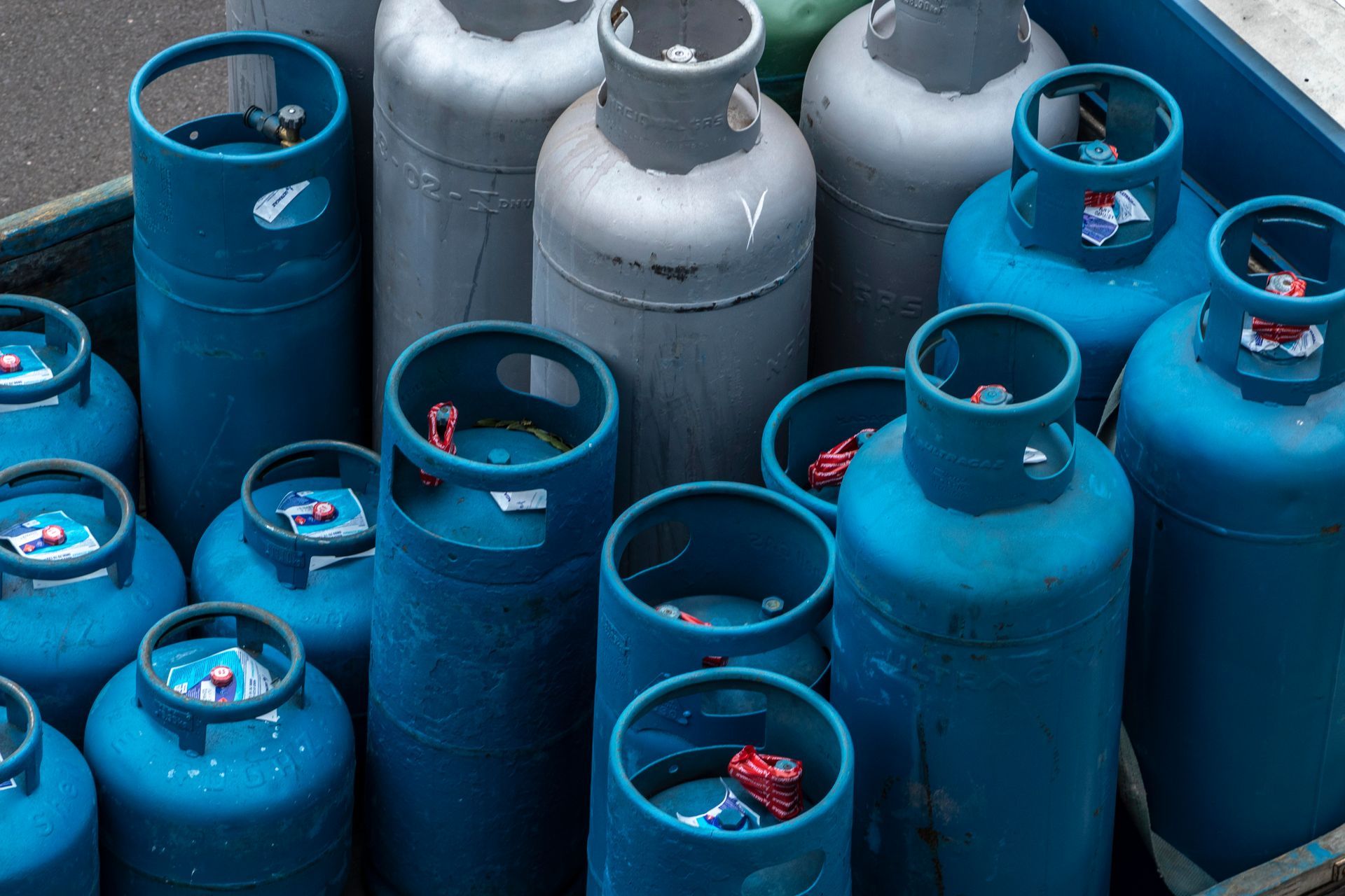 Tanques de propano azules en grupo, algunos grises, válvulas rojas visibles, al aire libre.