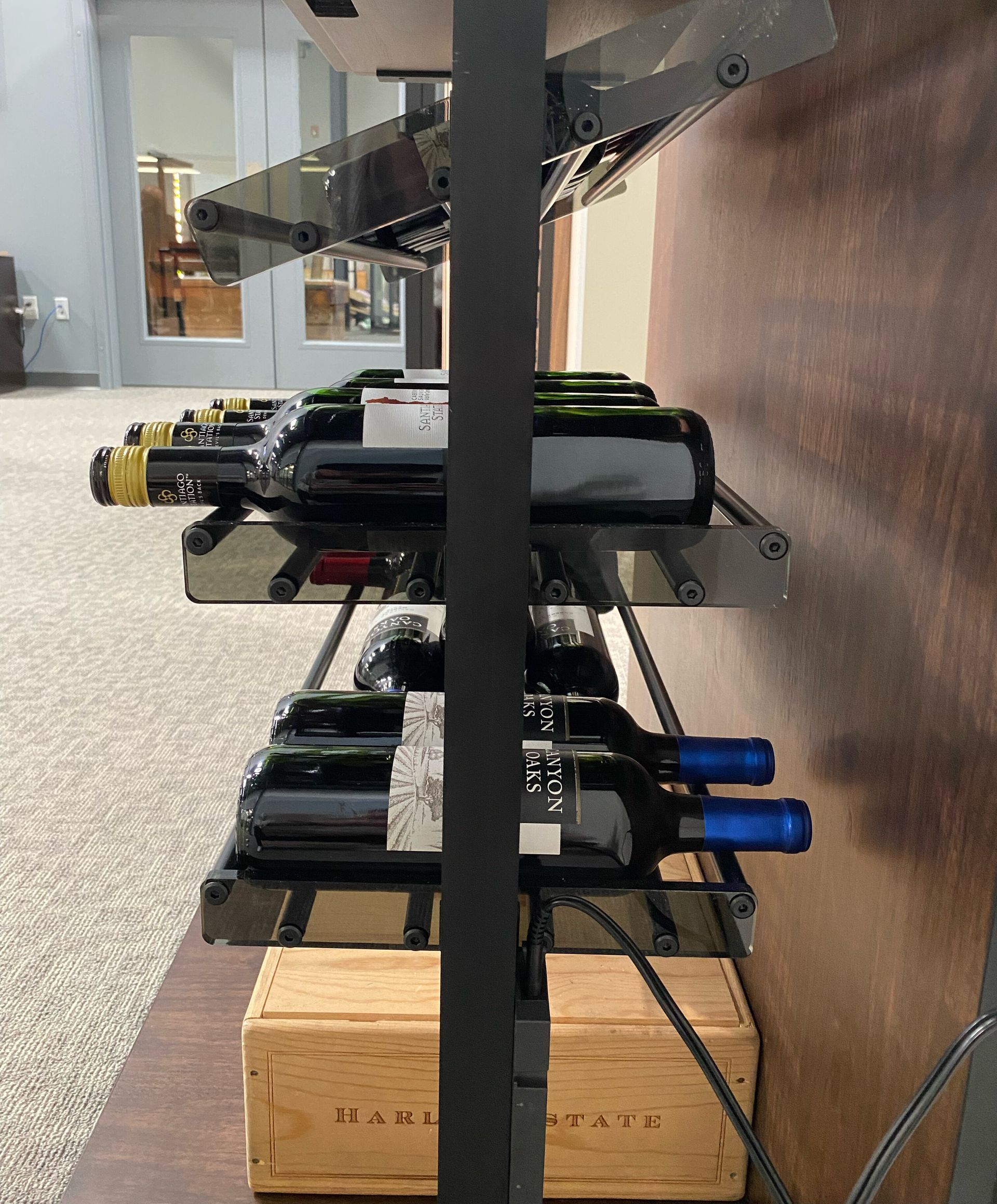 Black wine rack with bottles on glass shelves; wooden box below.
