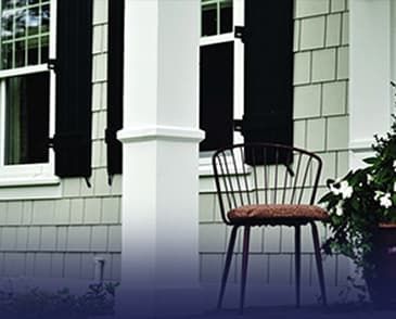 A chair is sitting on the porch of a house with black shutters.