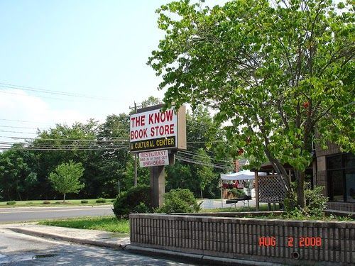 A sign for The Know Book Store and Cultural Center stands by a brick wall and a tree under a bright blue sky.