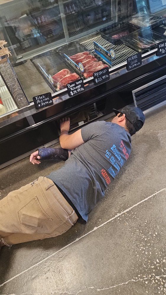 A man is laying on the floor in front of a meat display case in a butcher shop.