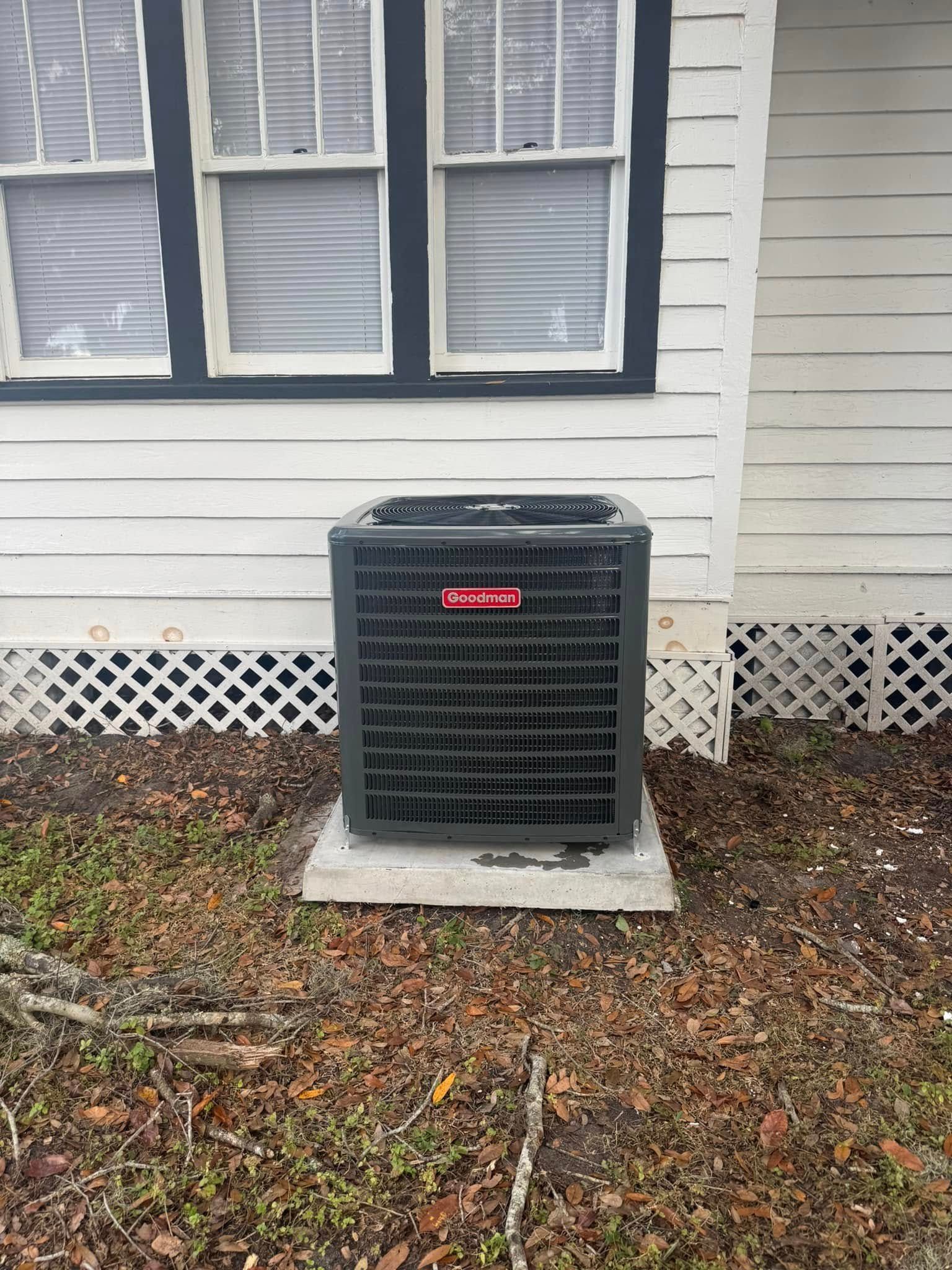 A black air conditioner is sitting in front of a white house.