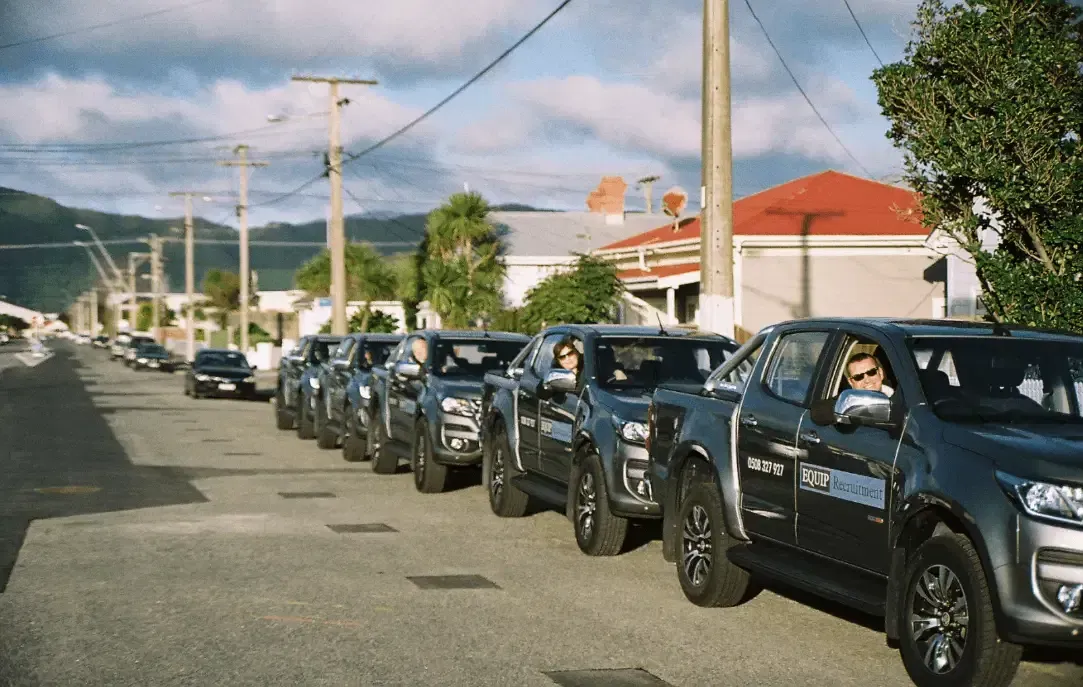 Line of dark pickup trucks driving down a street in a residential neighborhood under a blue sky
