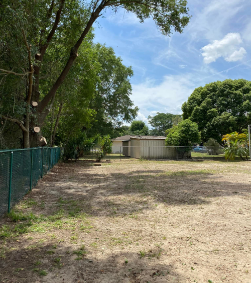 Empty dirt yard with green fence and trees under a partly cloudy sky.