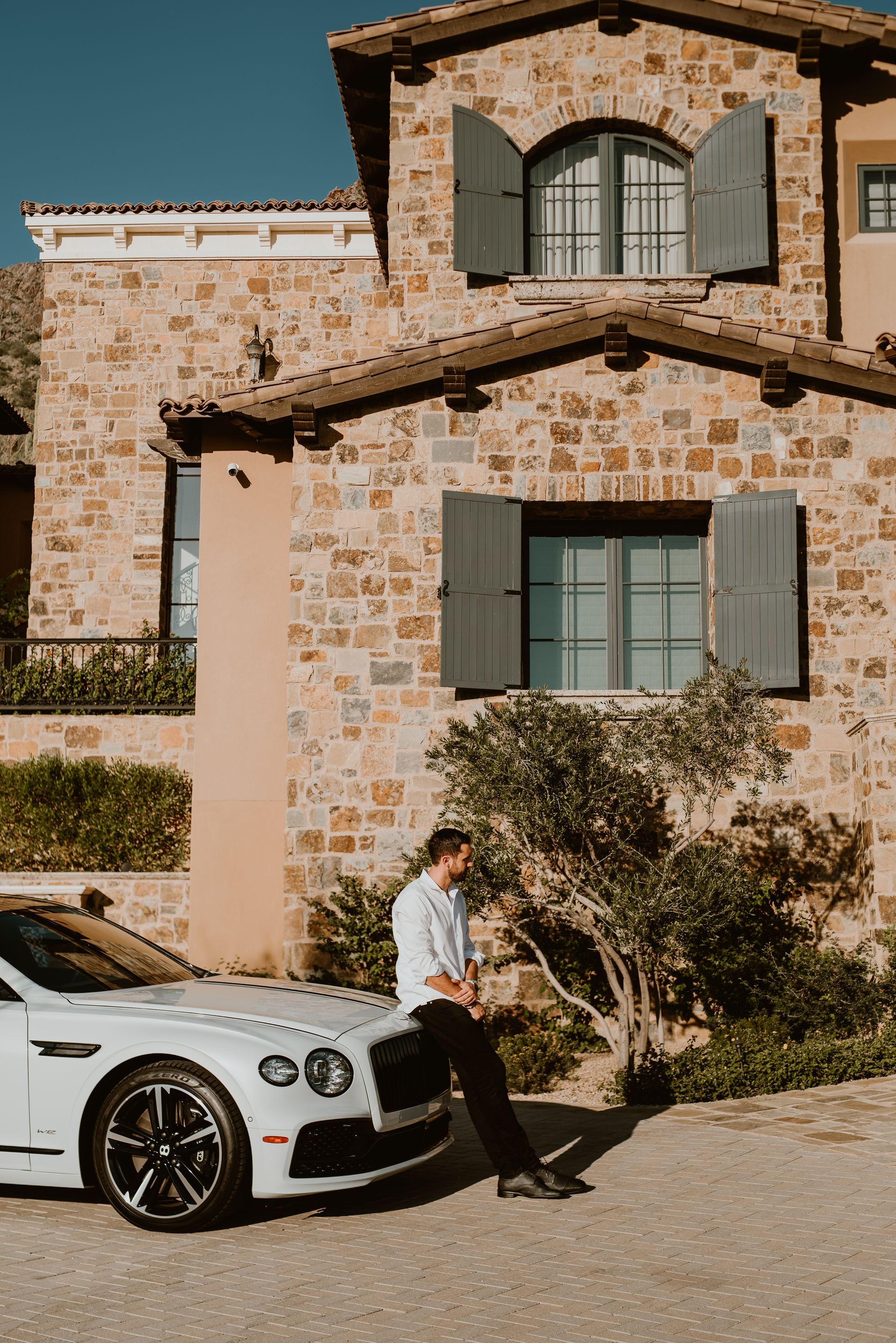 A man is sitting on the hood of a white car in front of a stone building.