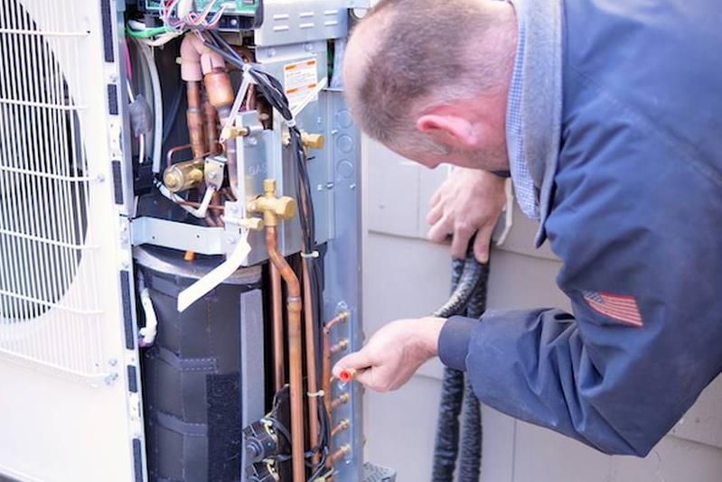 Technician in blue uniform repairs air conditioner unit outdoors, using a tool on copper pipes.