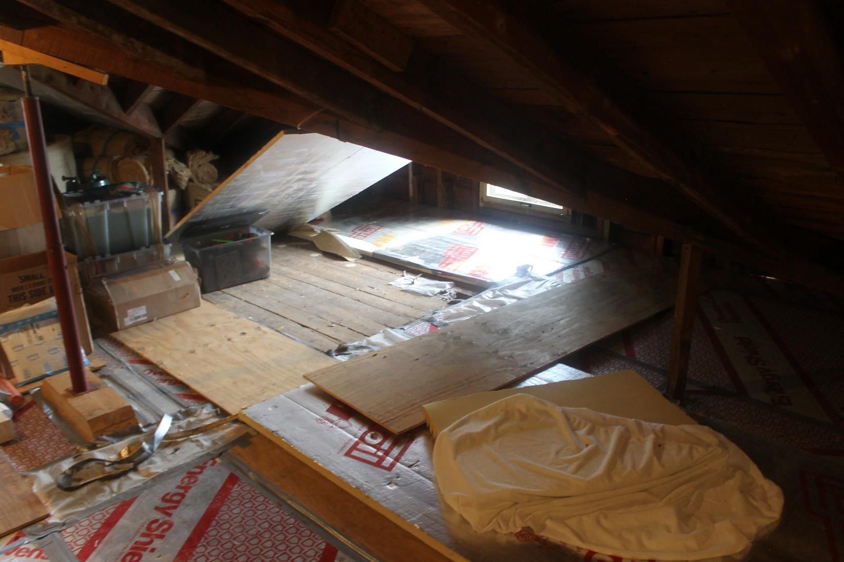 Attic interior with boards on insulation, bright light from an opening, and stored items.