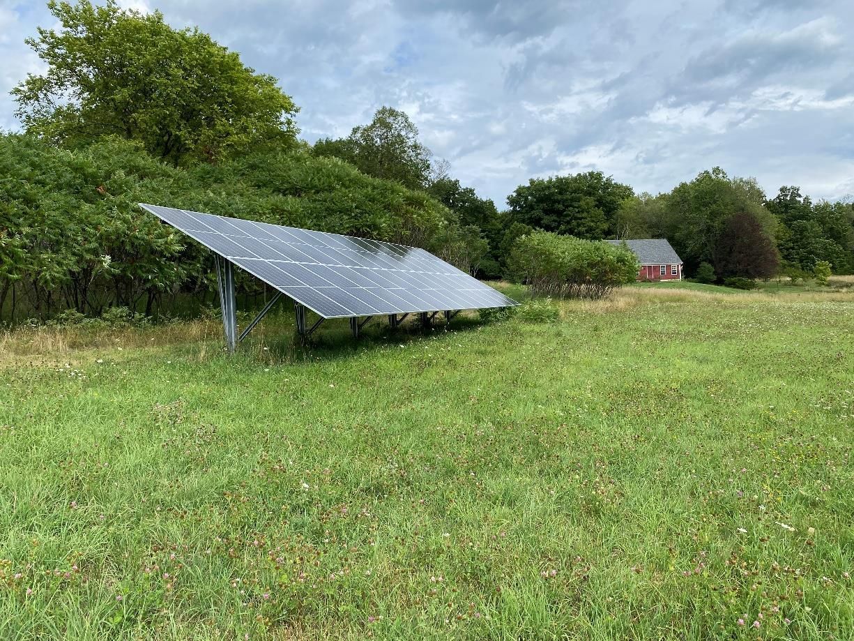 Solar panel in a grassy field, with trees and a red house in the background under a cloudy sky.