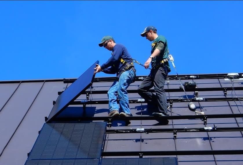 Two workers installing solar panels on a rooftop under a clear, blue sky.