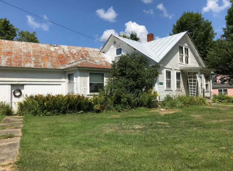 White, weathered house with overgrown yard, rusty roof, and a bright blue sky.