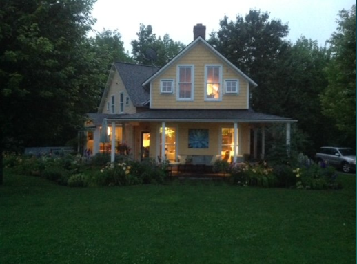 Yellow house at dusk with porch lights on, surrounded by greenery and a green lawn.