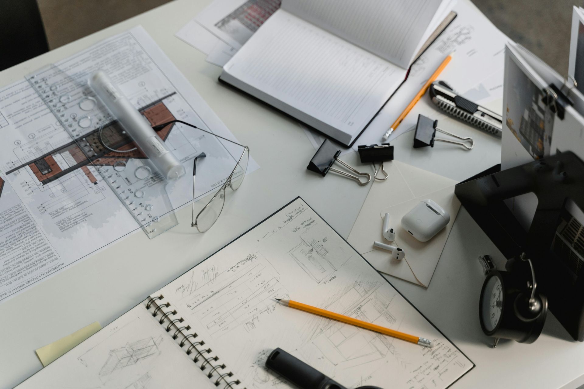Desk with architectural plans, notebooks, and stationery; pencils, a clock, and earbuds are also present.