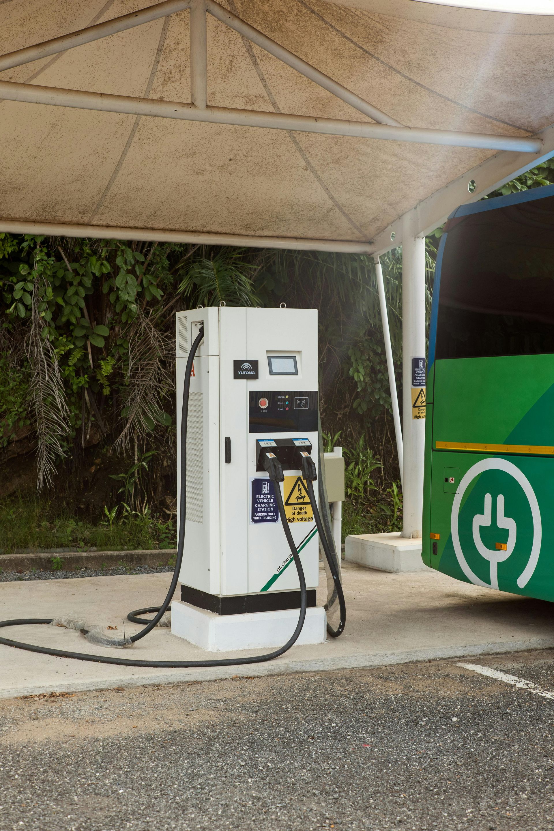 Electric bus charging at a station. White charger, green bus, concrete pad, covered structure, outdoor setting.