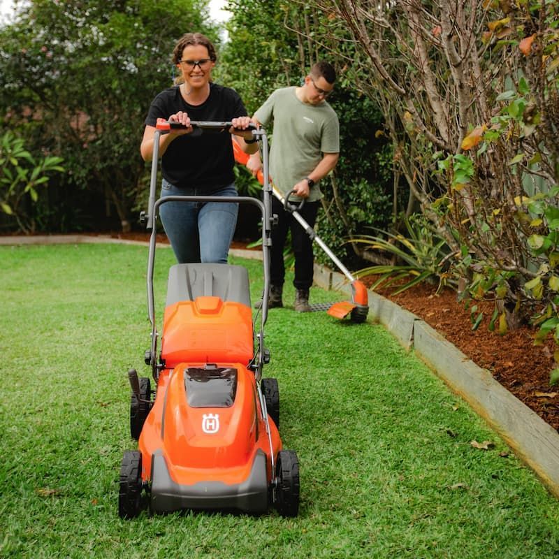 A Woman Is Pushing An Orange Husqvarna Lawn Mower — Mid Coast Mowers In Taree, NSW