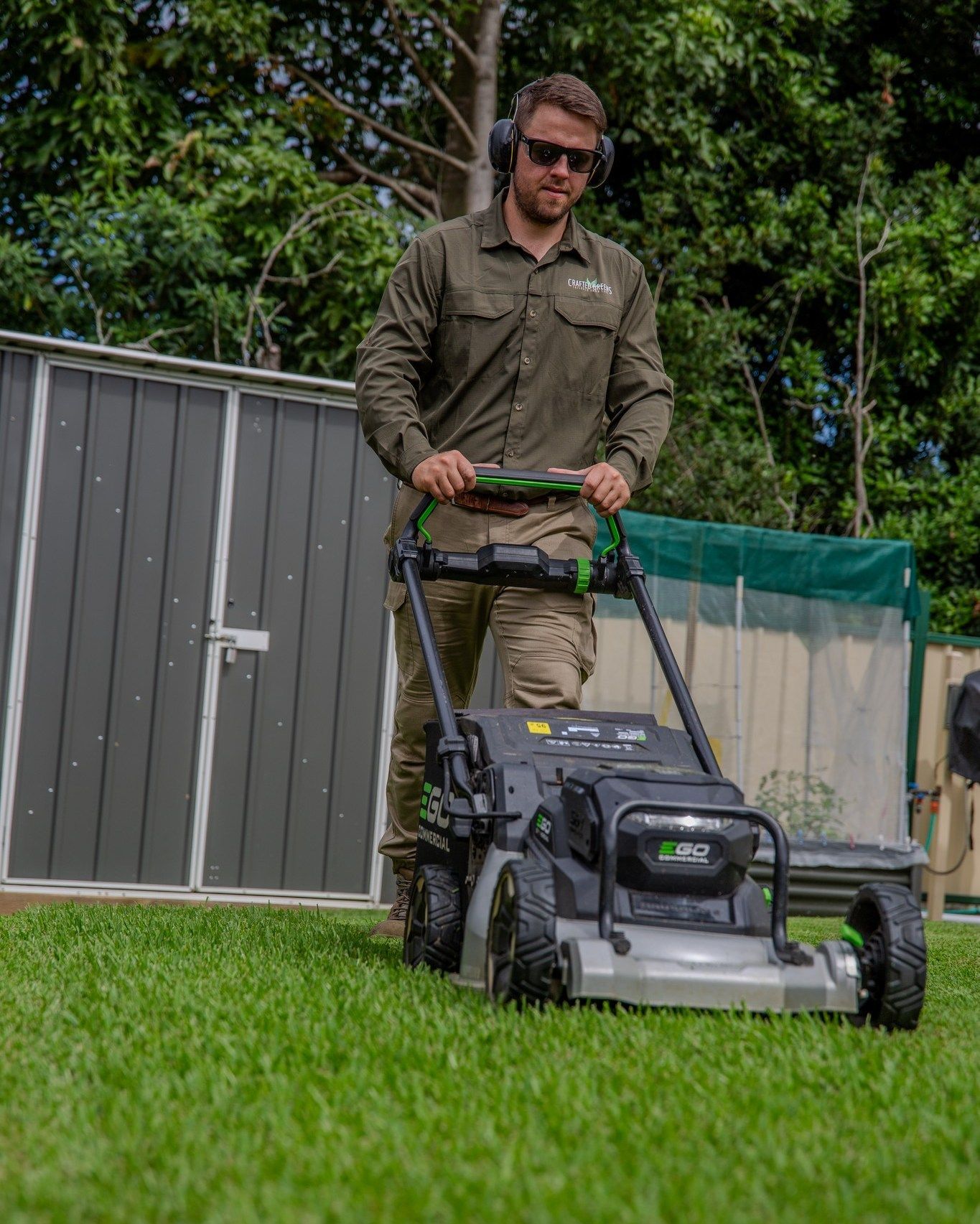 Man mowing a lawn with a push mower. He wears ear protection and sunglasses — Mow & Mulch Lawn & Garden Maintenance In Soldiers Point, NSW
