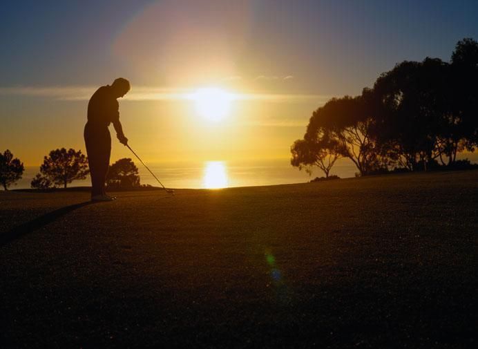 Golfer silhouetted against a setting sun over ocean, about to swing club on a green.
