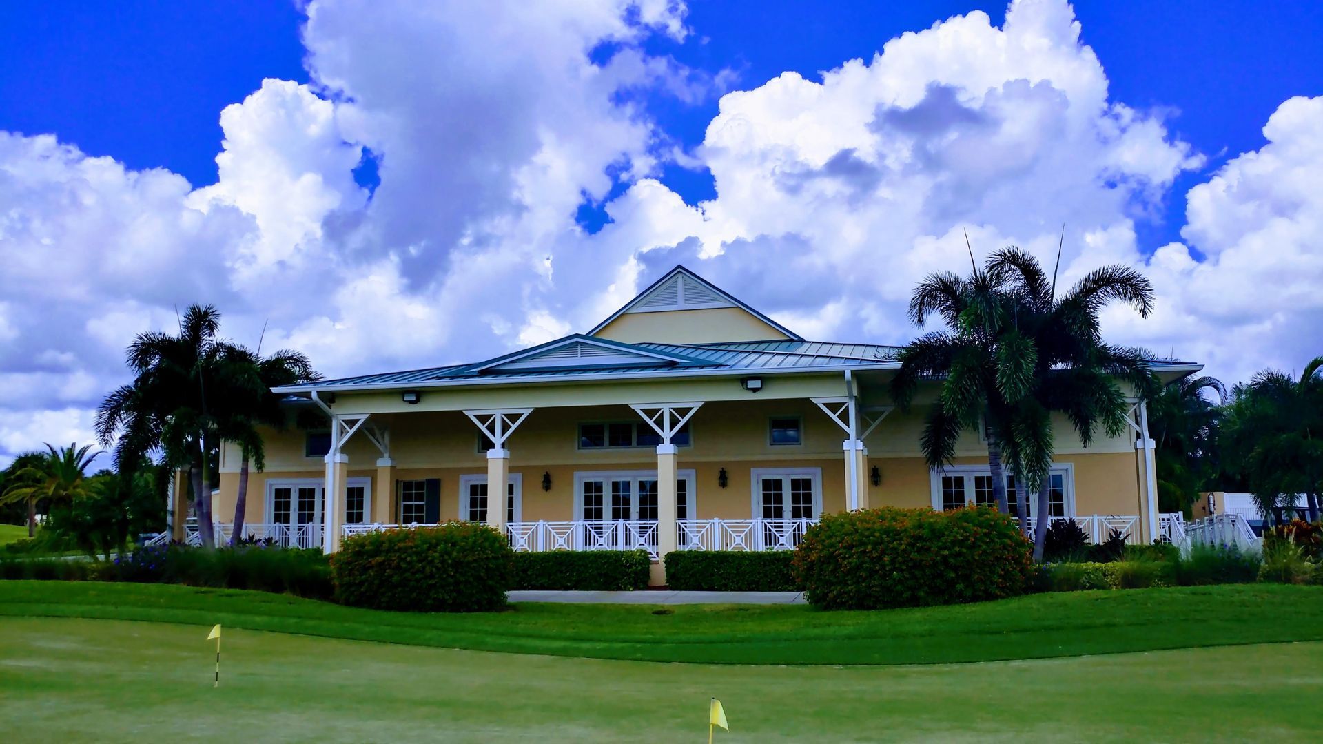 Yellow building with white accents, palm trees, and green lawn under a partly cloudy blue sky.