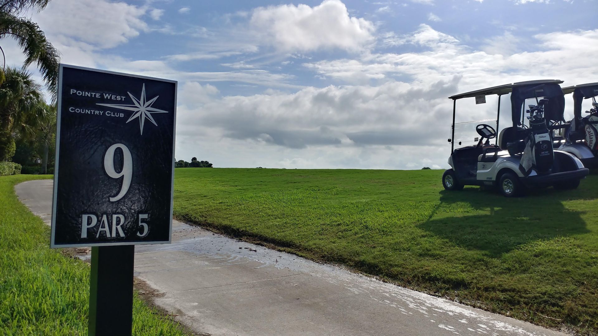 Golf course sign for hole 9, a par 5, with golf carts parked on a green fairway under a cloudy sky.