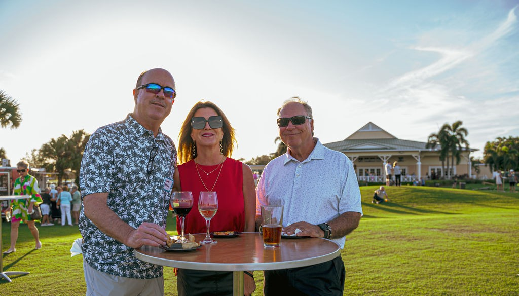 Three people at outdoor event, drinking by a table. Sunny day, golf course in background.