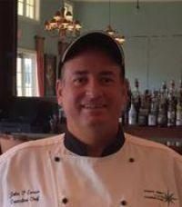 Chef in a white coat and black hat smiles in a restaurant setting.