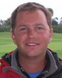 Man smiling, wearing a red jacket and blue shirt, outdoors on a golf course.