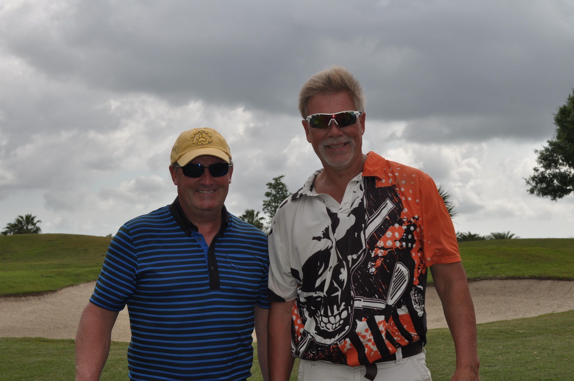 Two men in sunglasses on a golf course, one in a striped shirt, the other in a patterned shirt.