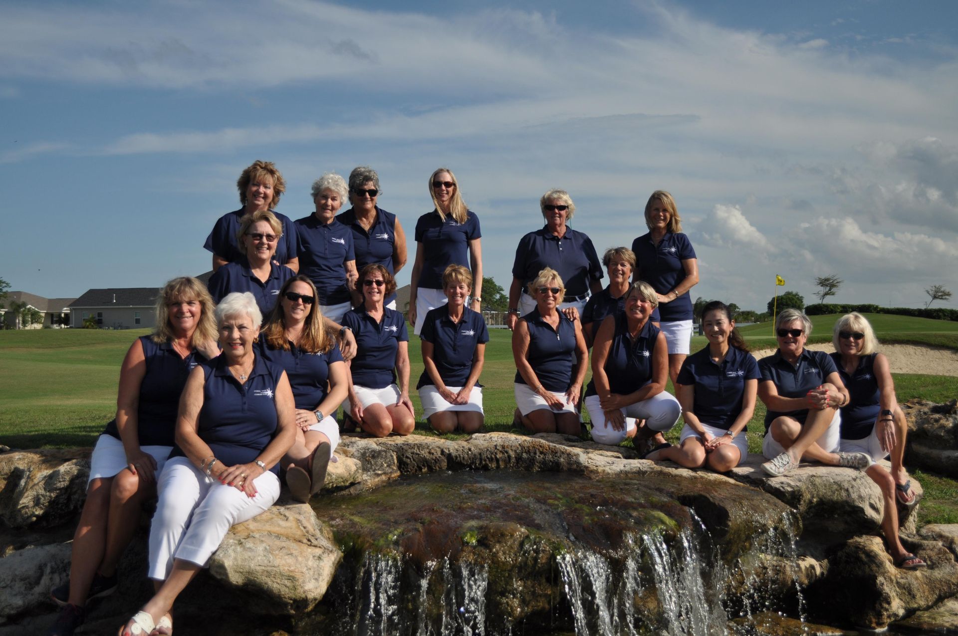 Group of women in navy shirts and white shorts posing near a small waterfall on a golf course.
