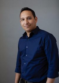 Man in a blue button-up shirt poses in front of a gray background, smiling slightly.