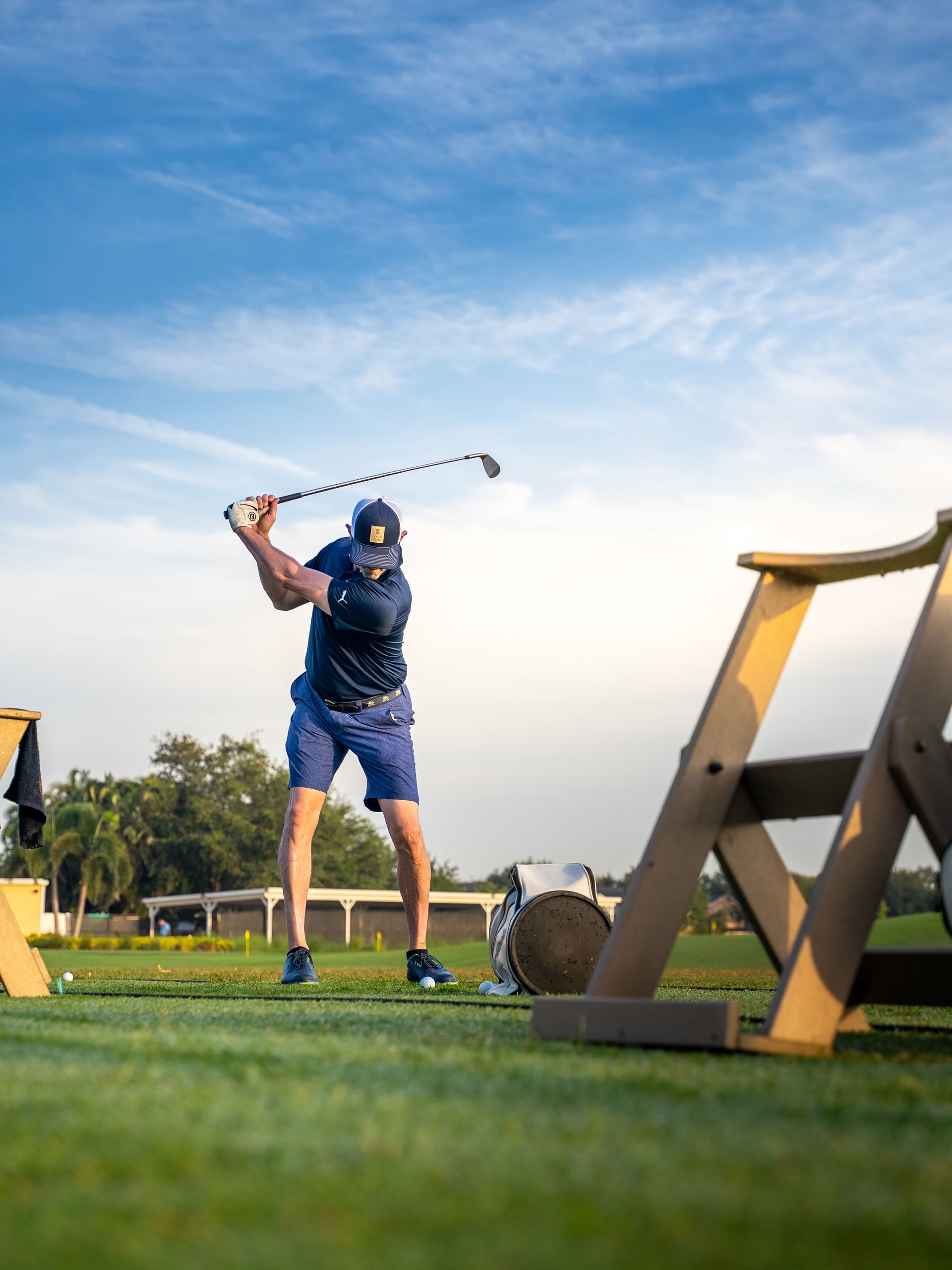 Man swinging a golf club on a green course, with blue shorts and shirt. Wooden structures and blue sky in background.