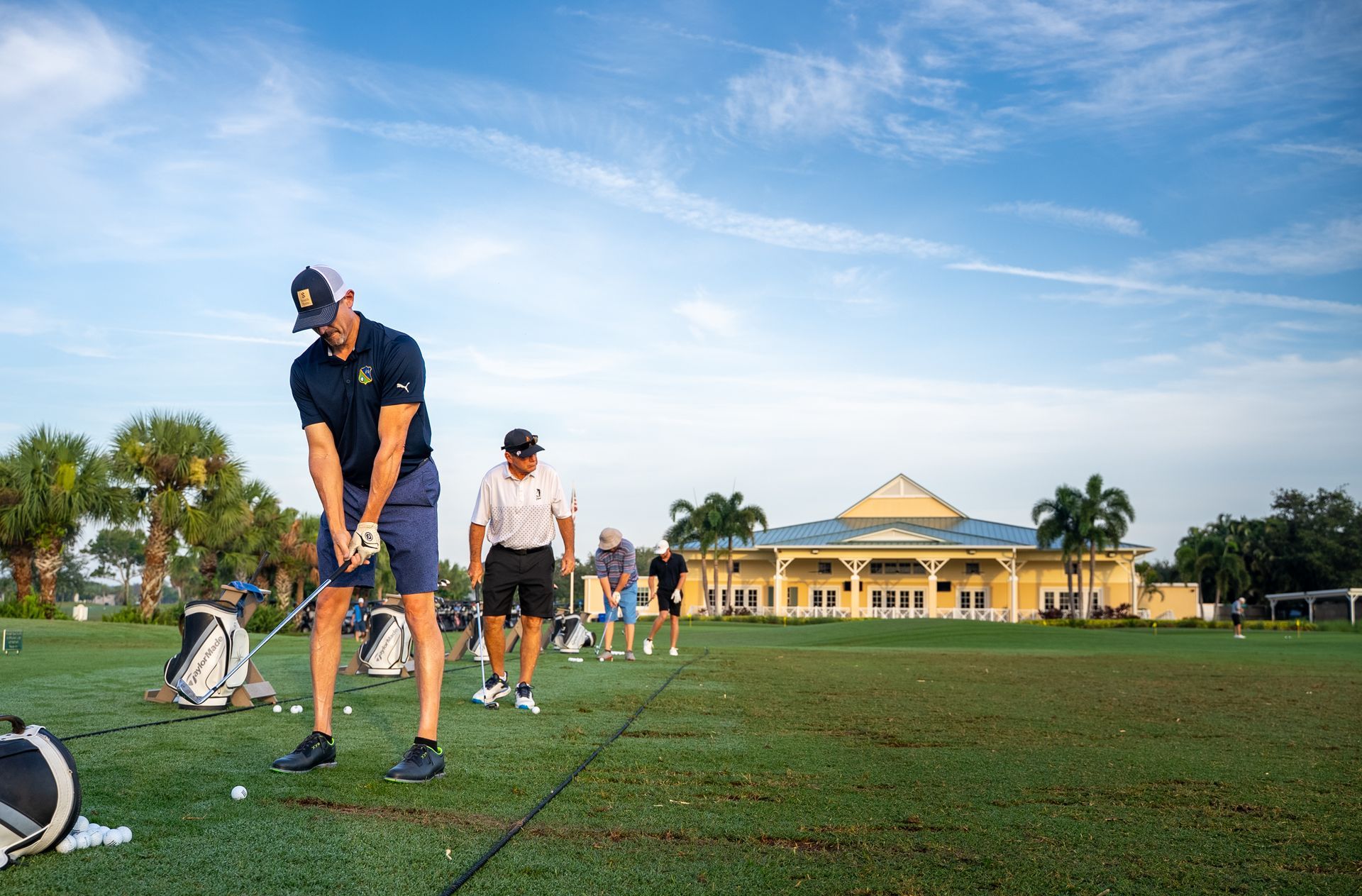 People practicing golf on a green field, a large building in the background. Sunny day.