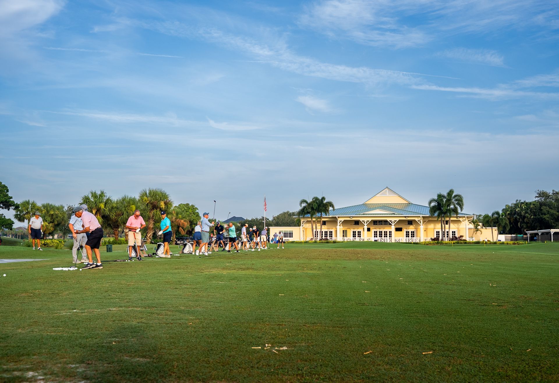People on a golf course practicing near a light-colored building with a blue sky background.
