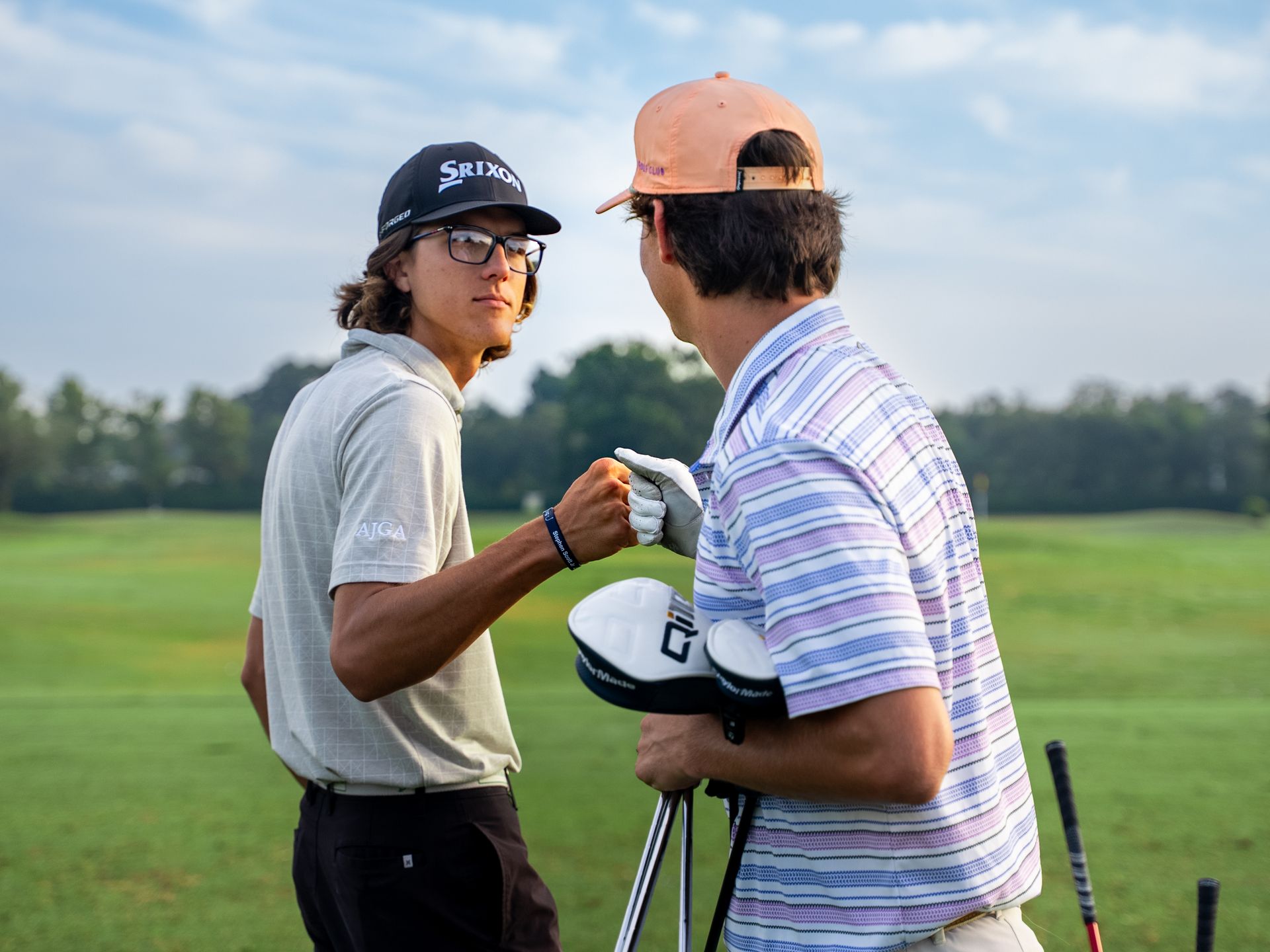 Two golfers bump fists on a green. One wears a black cap and glasses, the other a peach cap. Golf clubs are present.