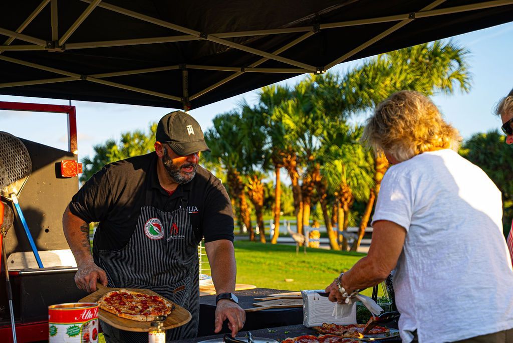 Man serving pizza to a woman at an outdoor event near palm trees.