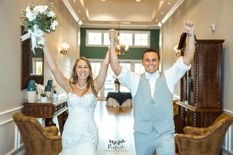 Newlywed couple, arms raised in triumph, walking down a hall. Bride in white dress, groom in vest.
