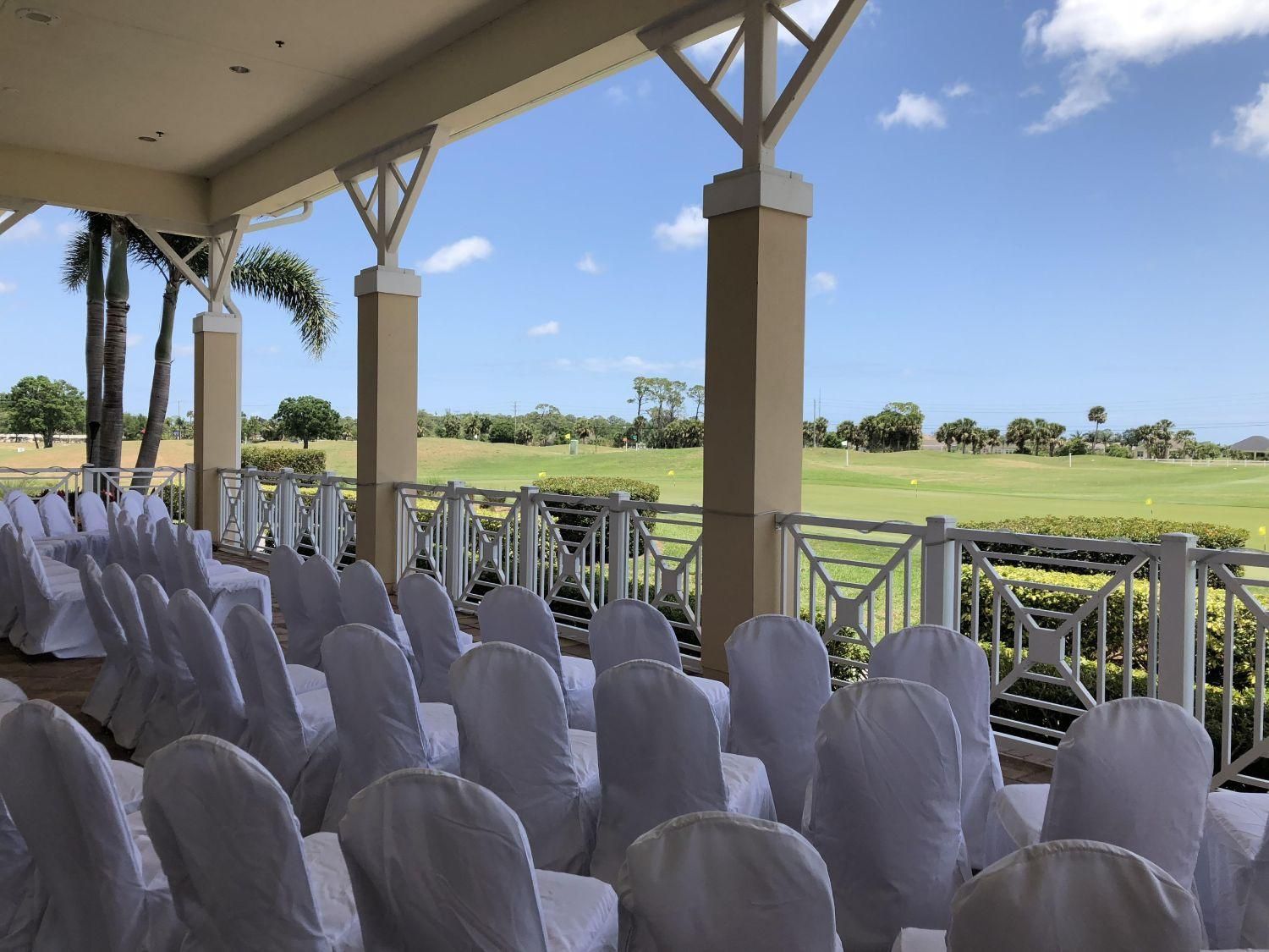 Rows of white-covered chairs arranged on a covered porch, overlooking a green golf course under a blue sky.
