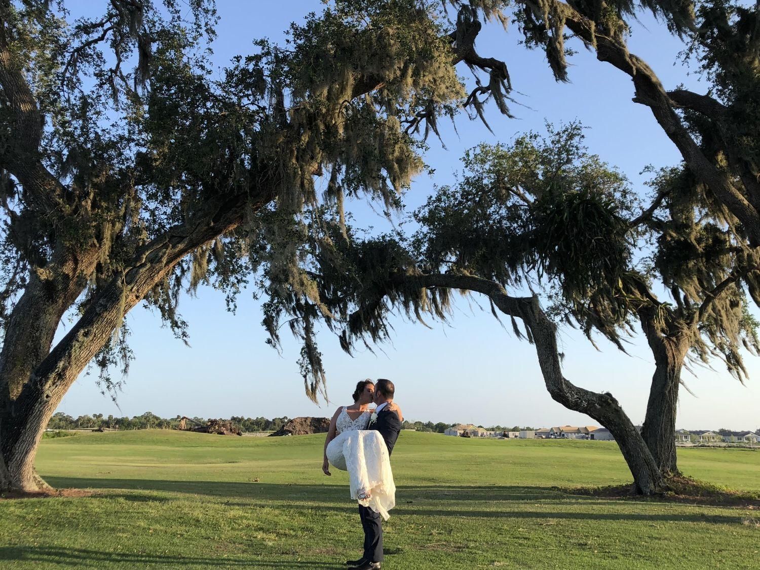 Man carrying a person in a wedding dress under large trees on a golf course.