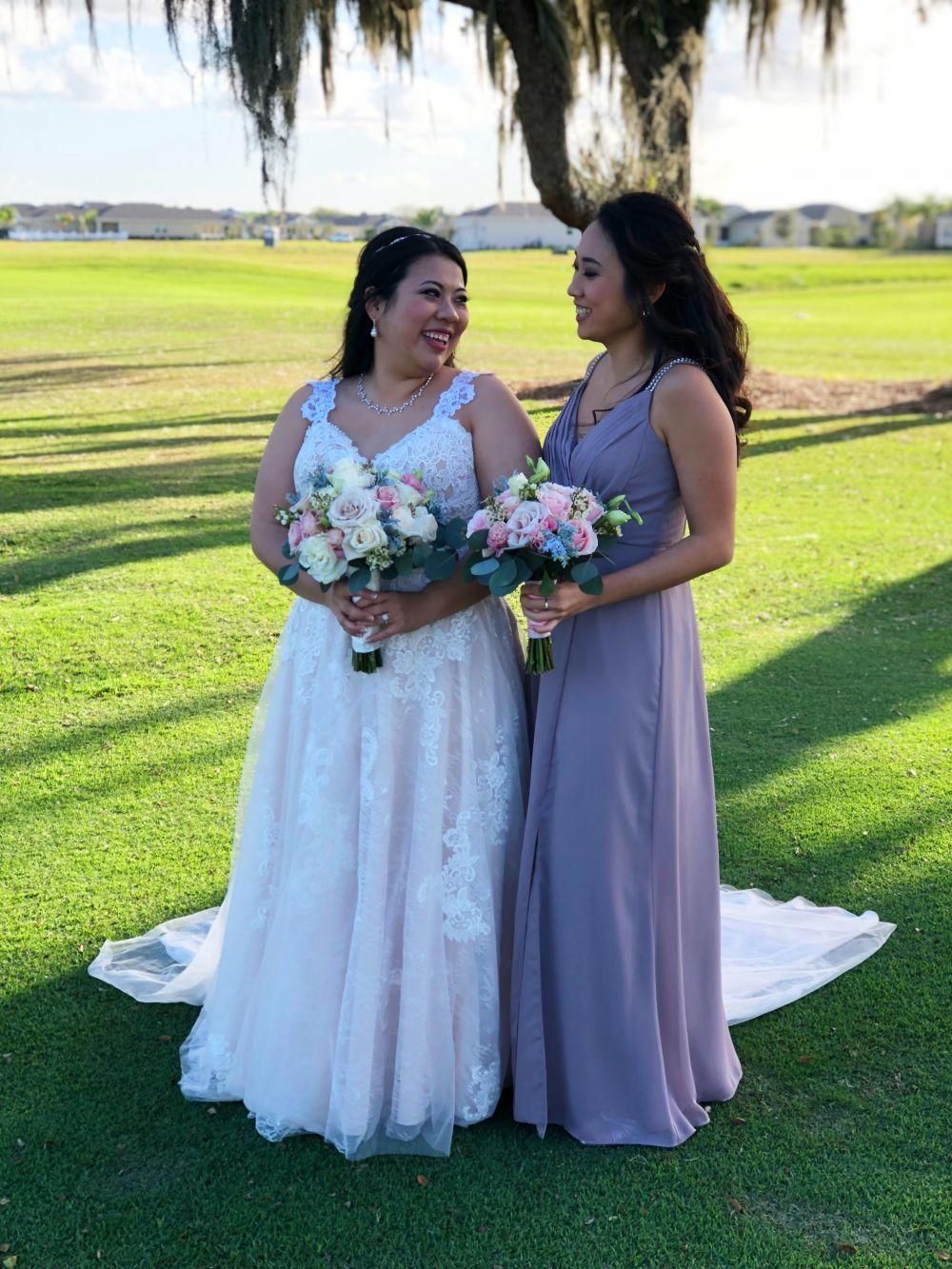 Bride and bridesmaid holding bouquets, smiling at each other on a golf course. Bride in white dress, bridesmaid in purple.