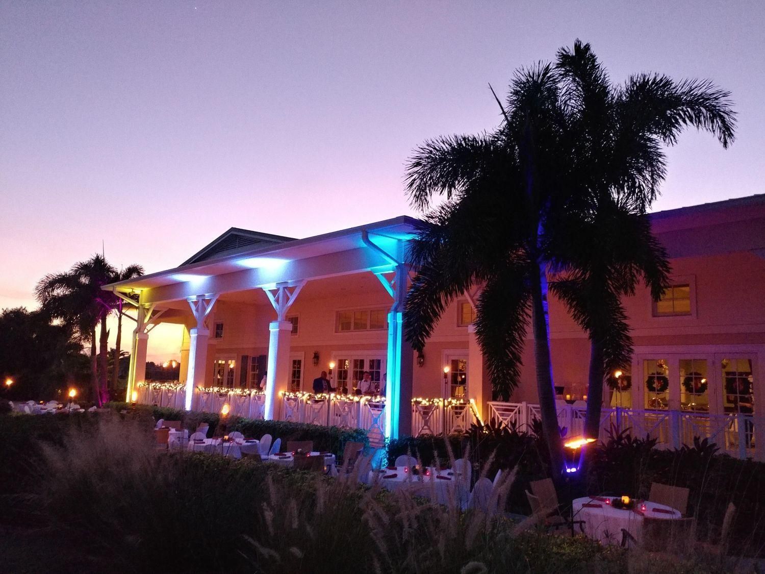 Building illuminated in blue, yellow, and white with palm trees, set at dusk. Tables set for an outdoor event.
