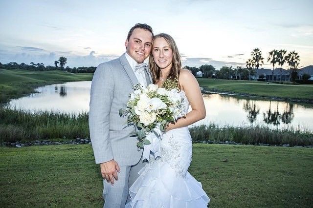 Newly married couple, man in gray suit and woman in white dress, pose outdoors by water, holding bouquet, smiles.