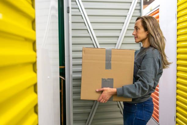 A woman wearing a grey sweater and jeans carries a box into a self storage solutions facility.