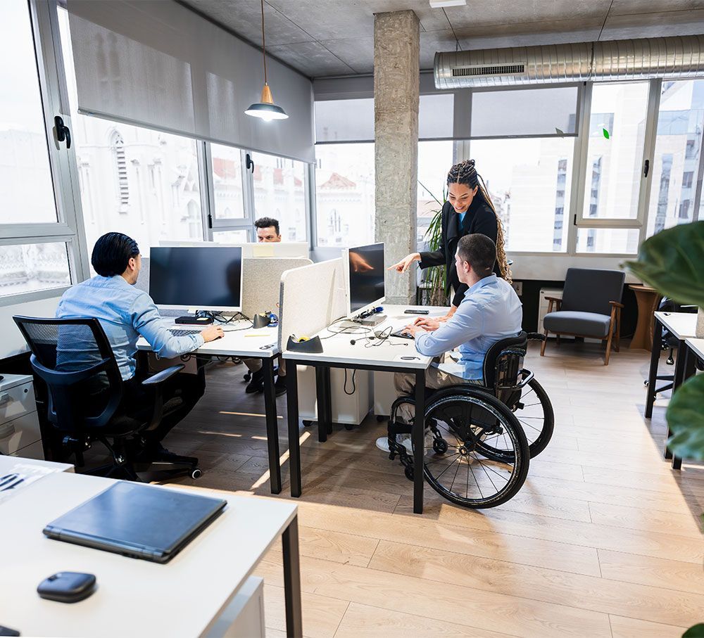 Office with three people at desks. One in a wheelchair. A woman points at a computer.