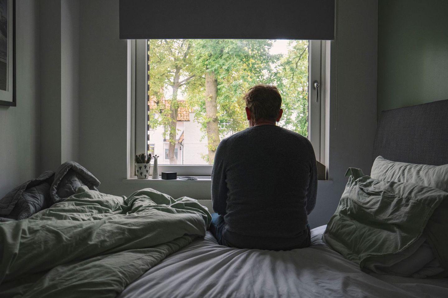 A man is sitting on a bed looking out of a window.