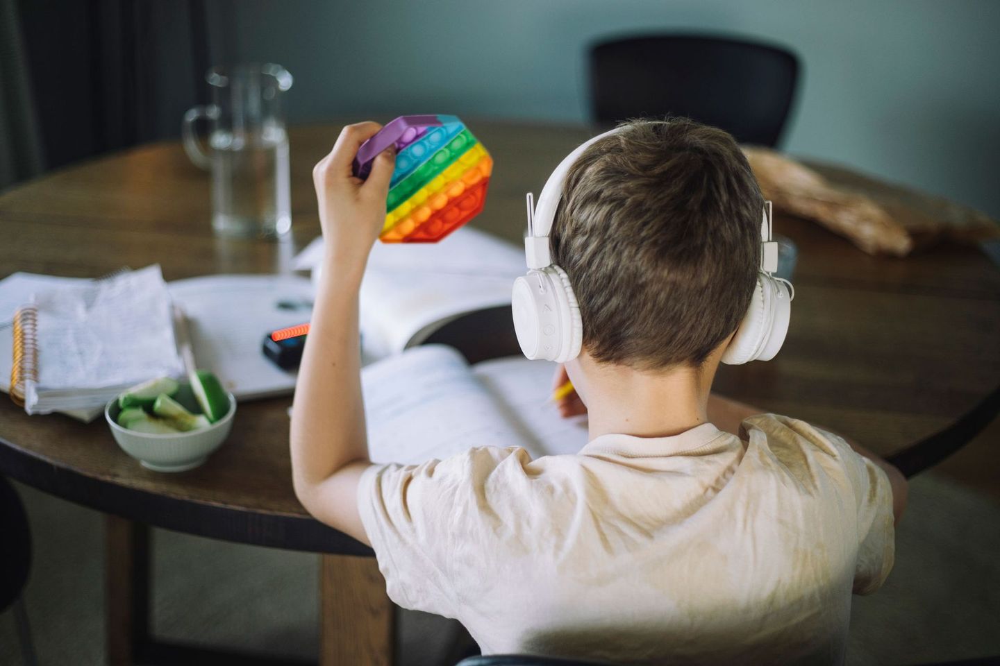 A young boy wearing headphones is sitting at a table holding a rainbow pop it.