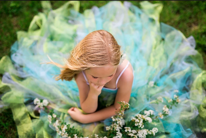 A little girl in a blue and green dress is sitting in the grass holding flowers.