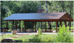 A large wooden pavilion with a metal roof in the middle of a park.