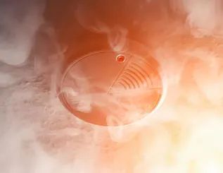 Smoke-filled room with a smoke detector on the ceiling; emitting red light.