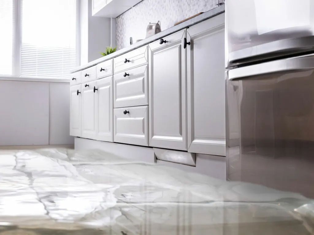 Kitchen with water flooding the floor, in front of white cabinets. Refrigerator on right.