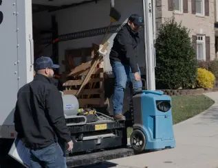 Movers loading equipment, including a blue machine, from a truck parked in a residential driveway.