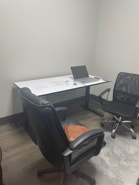 Small office with a desk, laptop, and two office chairs.  One chair has a brown seat.